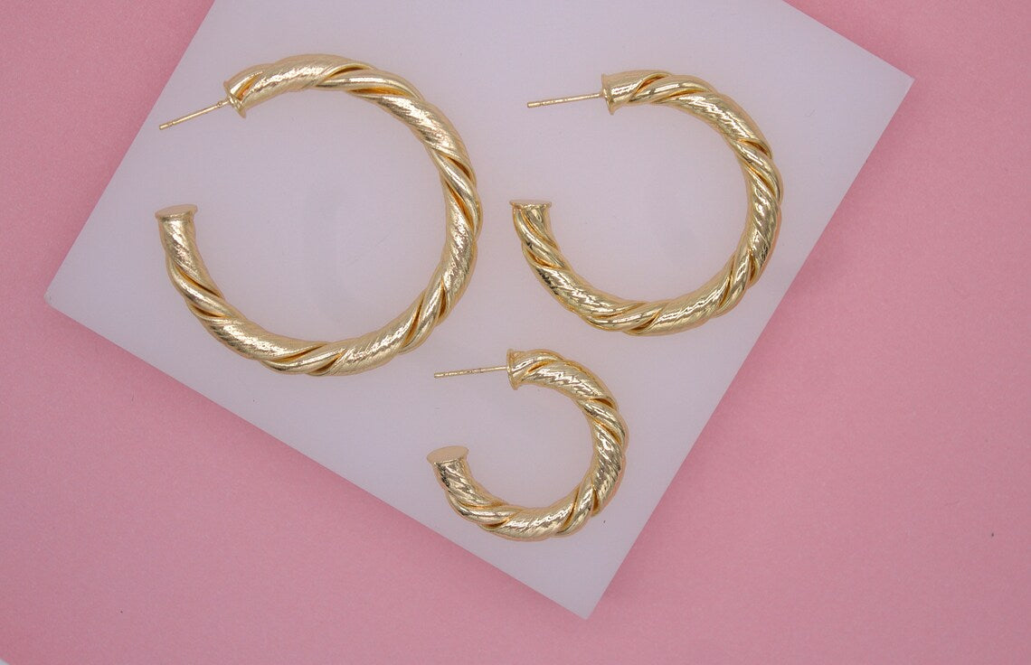 Three graduated sizes of textured gold hoop earrings rest on a white display stand against a pink background.