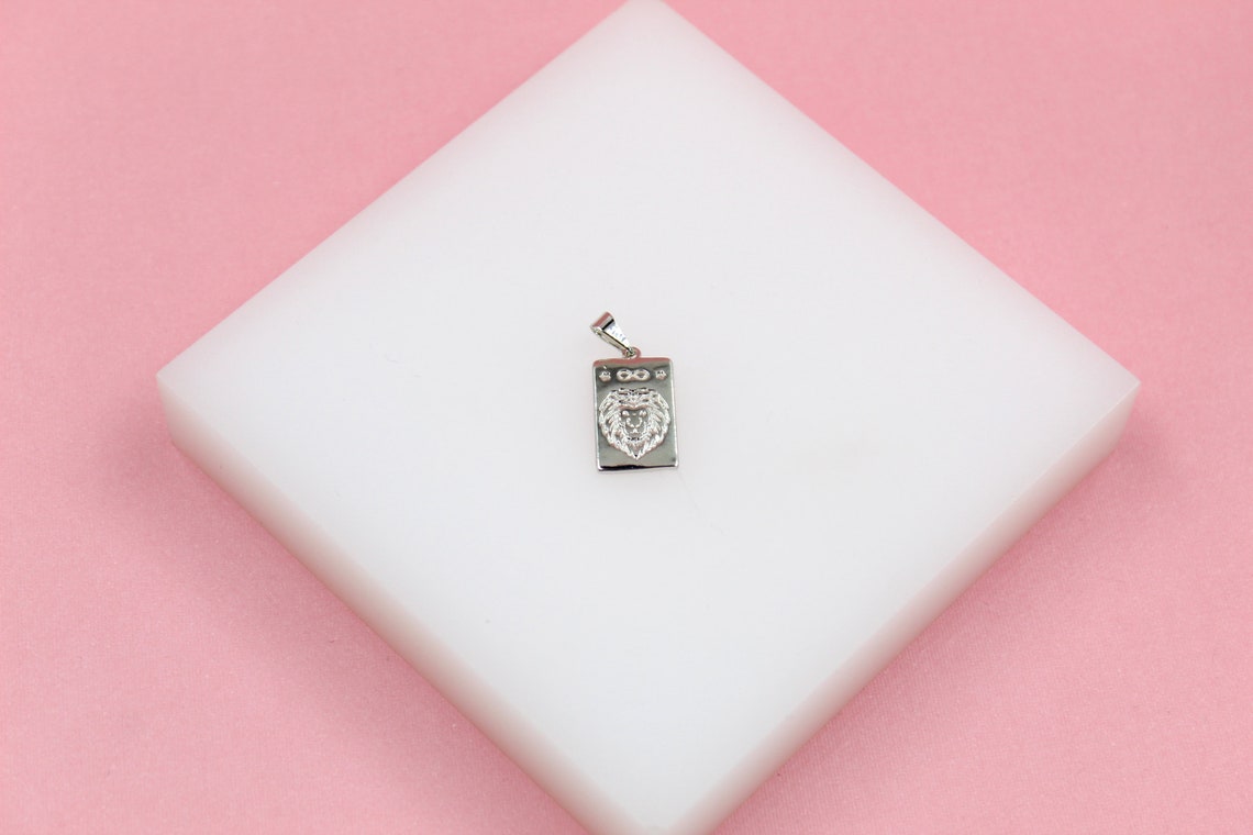 A silver rectangular lion pendant rests on a white display block against a pink background.