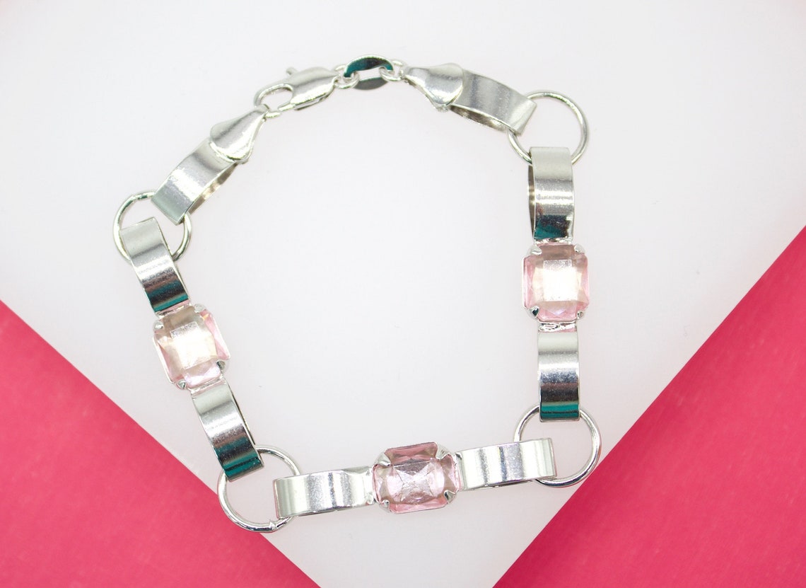 A silver link bracelet features alternating curved metallic sections and square light pink crystal stones against a pink and white background.