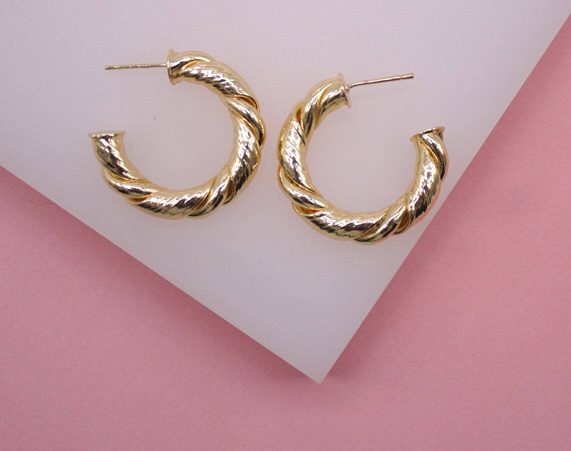 Two shiny gold twisted hoop earrings rest side by side on a white and pink display surface.