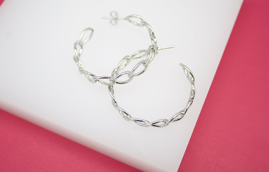 A pair of stylish silver chain link hoop earrings rests on a white display surface against a vibrant pink background.