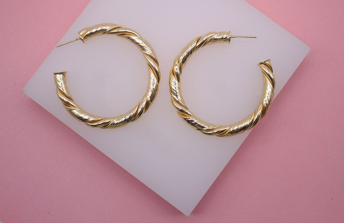A pair of textured gold hoop earrings rests neatly on a white display stand against a vibrant pink background.