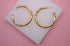 A pair of textured gold hoop earrings rests neatly on a white display stand against a vibrant pink background.
