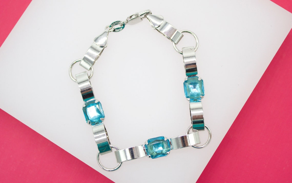 A silver link bracelet adorned with sparkling aquamarine gemstones rests on a white and pink surface.