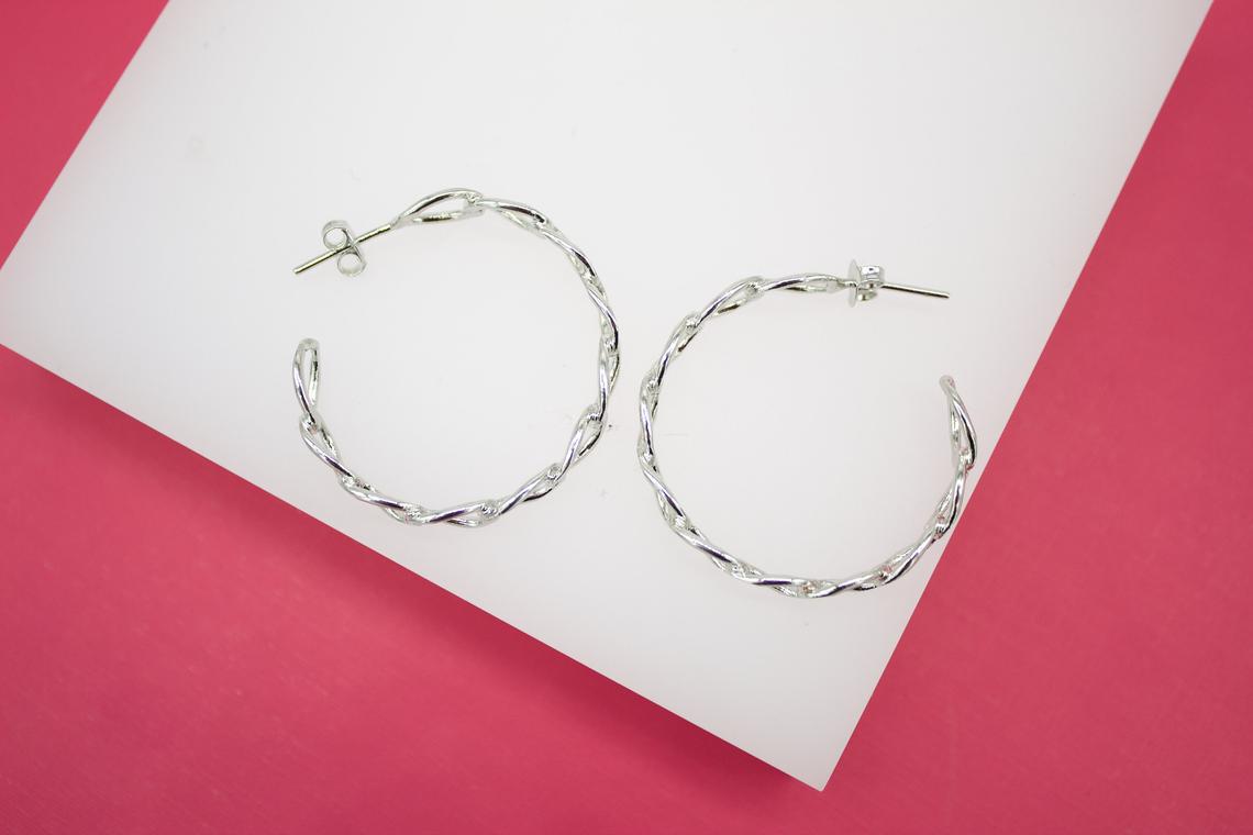 A pair of silver twisted hoop earrings rests on a white display surface against a vibrant pink background.