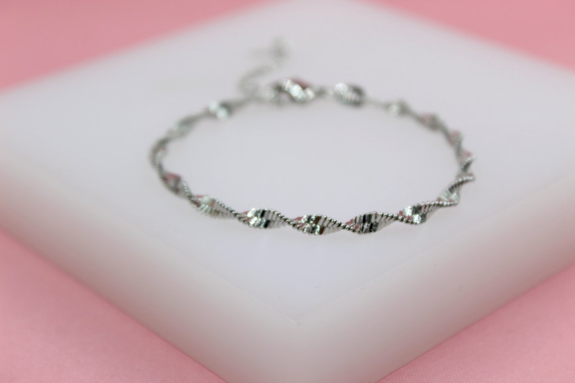 A delicate silver twisted chain bracelet rests on a white display block against a soft pink background.