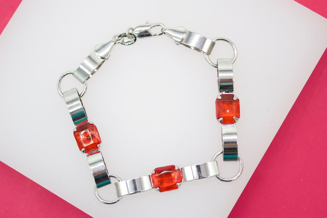 A silver link bracelet featuring vibrant orange gemstone stations rests on a white and pink background.