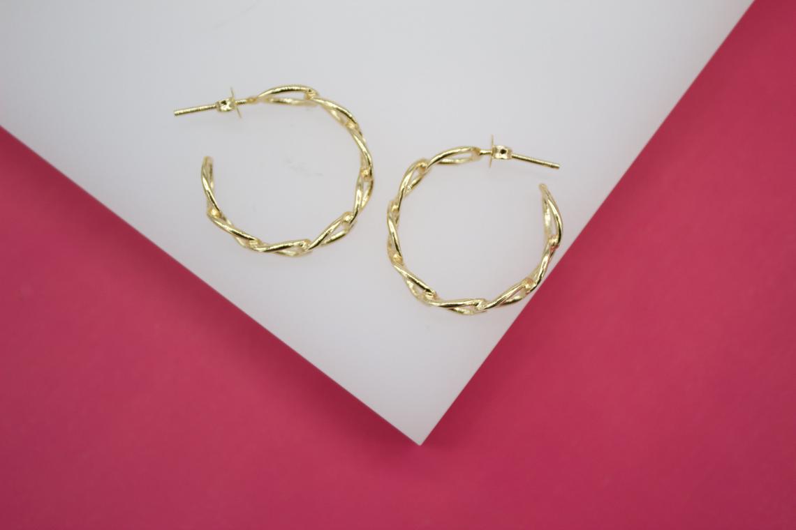 A pair of gold twist-textured hoop earrings rests on a white display against a pink background.