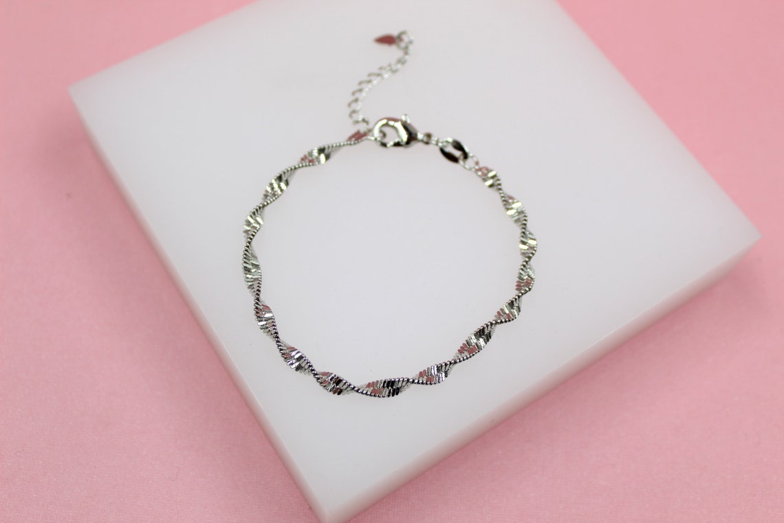 A delicate silver-plated twisted chain bracelet rests on a white display stand against a pink background.