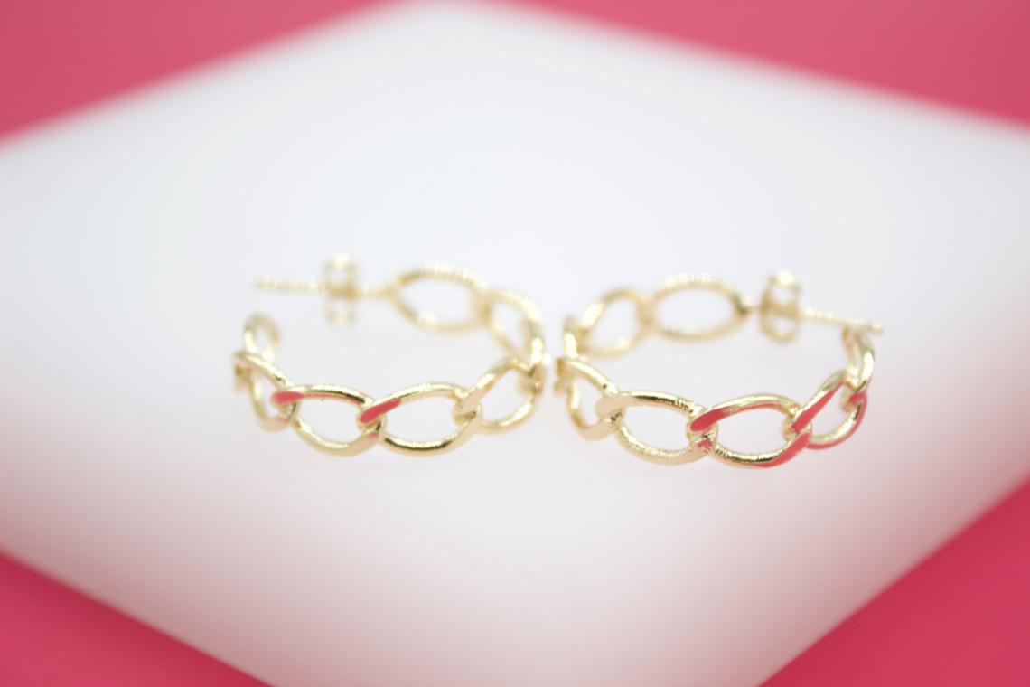 Two gold chain-link hoop earrings rest symmetrically on a white display surface against a bright pink background.