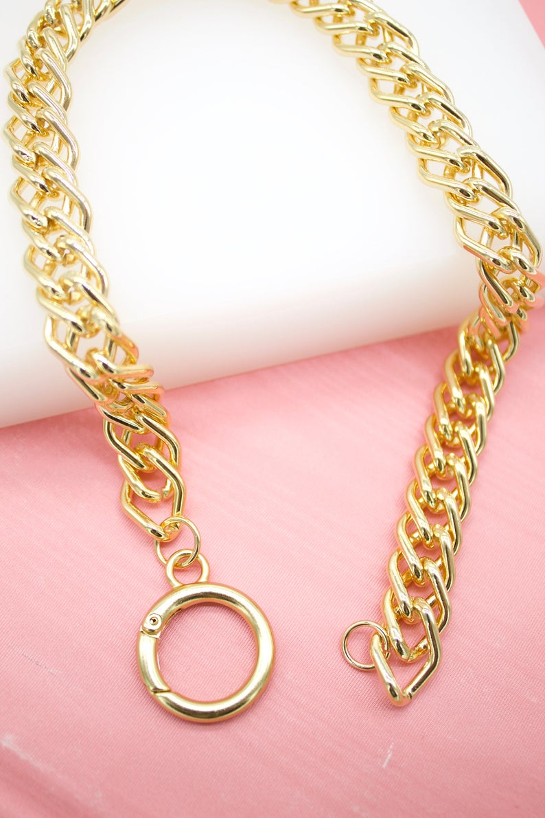 A thick, shiny gold chain necklace rests on a soft pink surface, featuring a distinct round spring ring clasp.