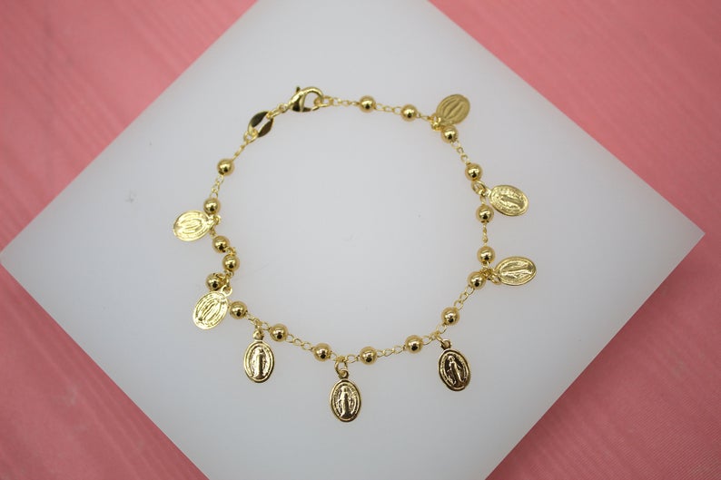 A gold-plated Catholic rosary bracelet featuring small bead accents and hanging Miraculous Medal charms rests on a white display surface.