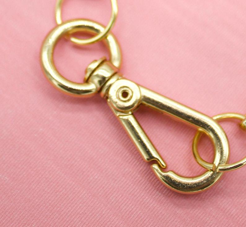 A gold-tone lobster clasp hangs against a pink background, securing jewelry jump rings.