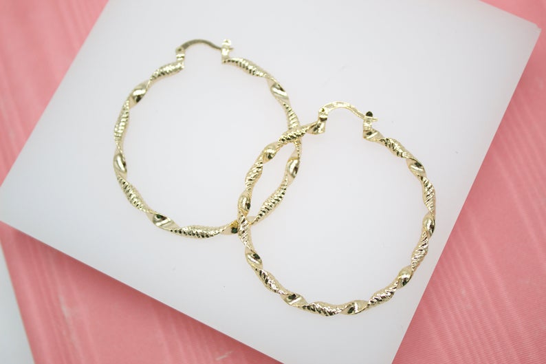 A pair of large, gold-plated twisted hoop earrings rests on a white display surface against a pink background.