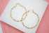 A pair of large, gold-plated twisted hoop earrings rests on a white display surface against a pink background.