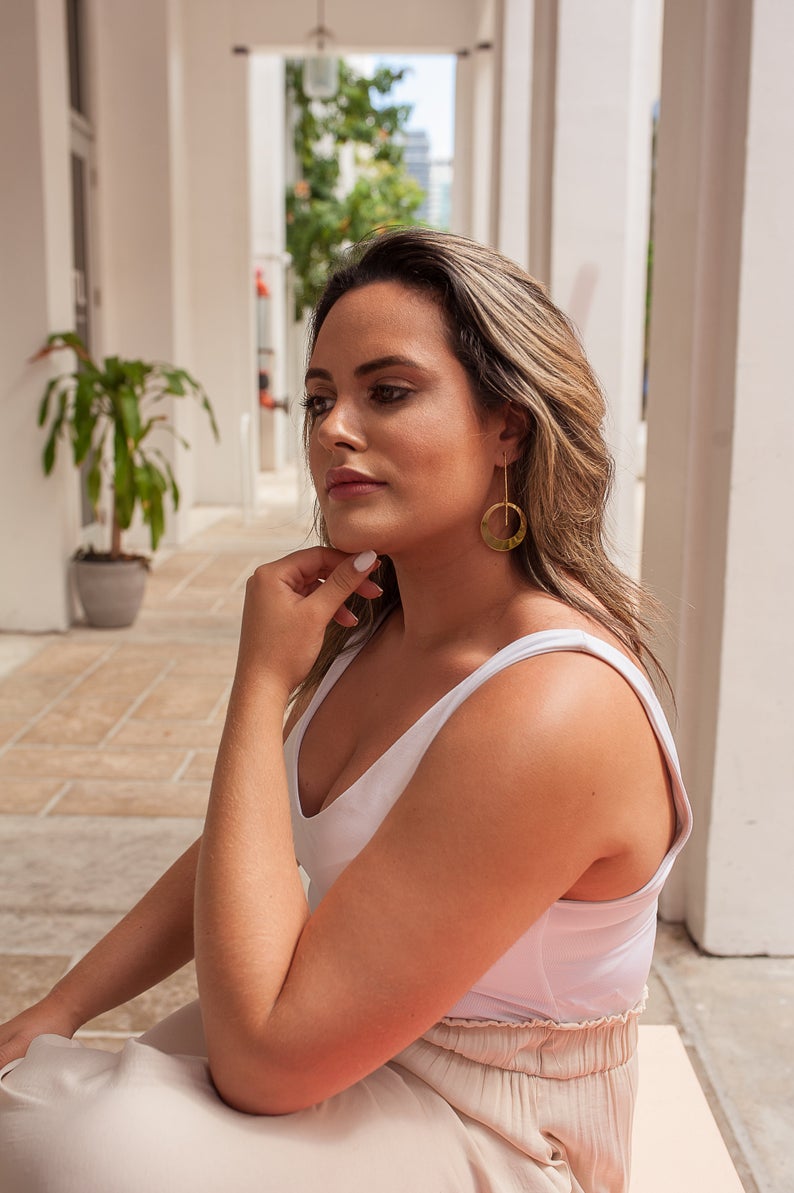 A stylish woman wearing large gold statement earrings rests her chin on her hand while posing gracefully in a sunlit architectural corridor.