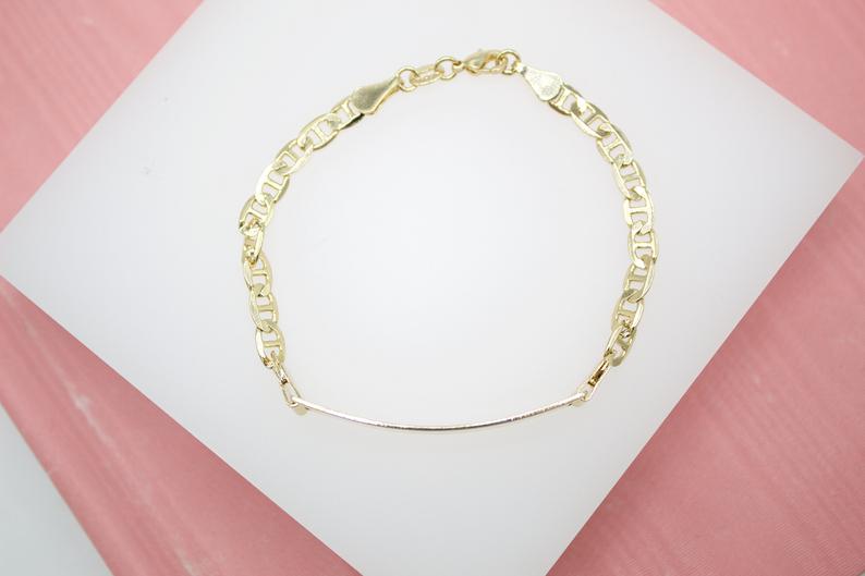 A gold mariner link chain bracelet with a curved ID bar rests on a white display against a pink background.