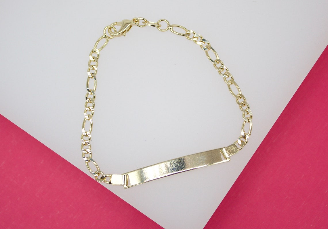 A shiny gold ID bar bracelet rests on a display surface featuring white and pink backgrounds.