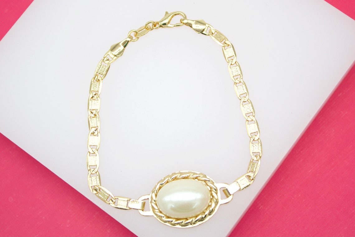 A gold link bracelet featuring a central oval faux pearl pendant rests on a white display surface against a pink background.