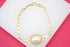 A gold link bracelet featuring a central oval faux pearl pendant rests on a white display surface against a pink background.