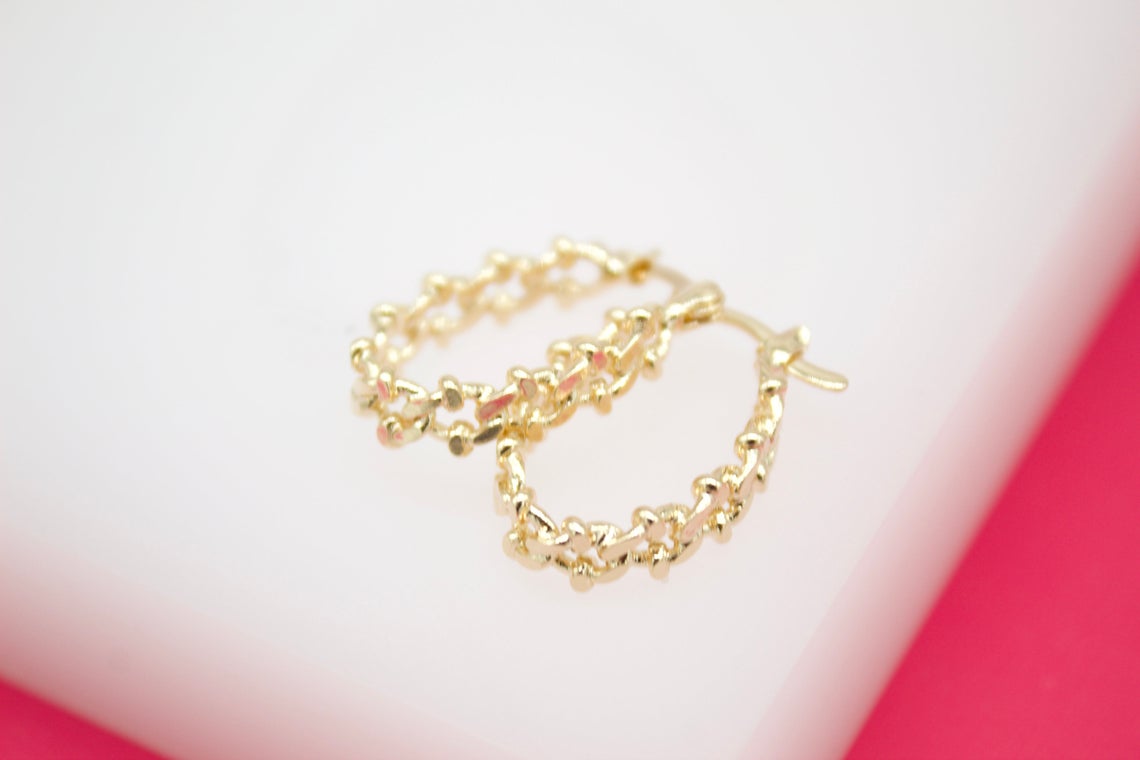 A pair of gold textured hoop earrings rest on a white surface against a bright pink background.