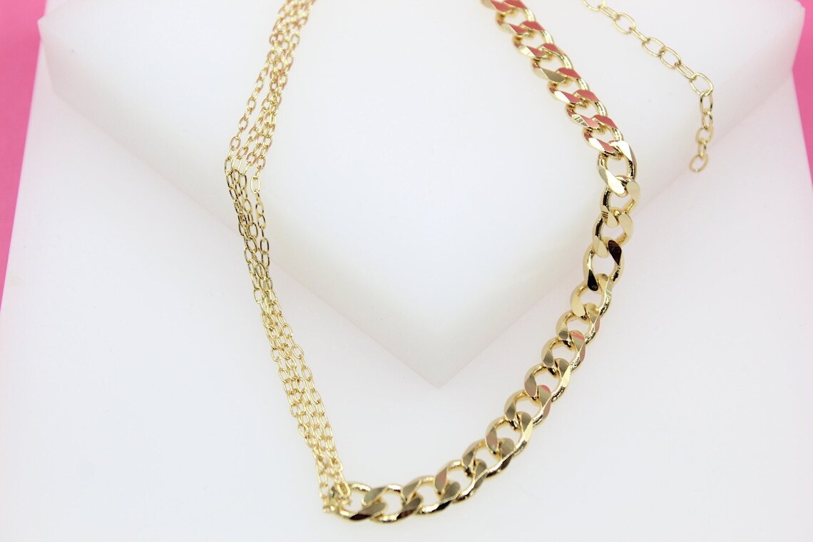 A gold layered choker necklace featuring a combination of fine delicate chains and a bold curb chain rests on a white display surface.