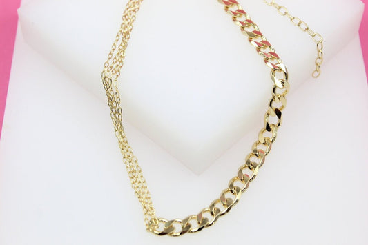 A gold layered choker necklace featuring a combination of fine delicate chains and a bold curb chain rests on a white display surface.