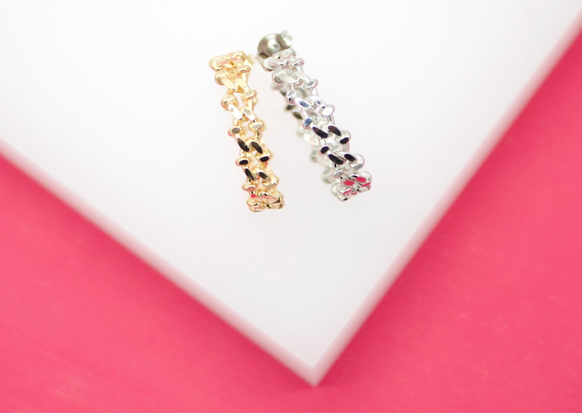 Two textured chain-link earrings in gold and silver rest on a white surface against a pink background.