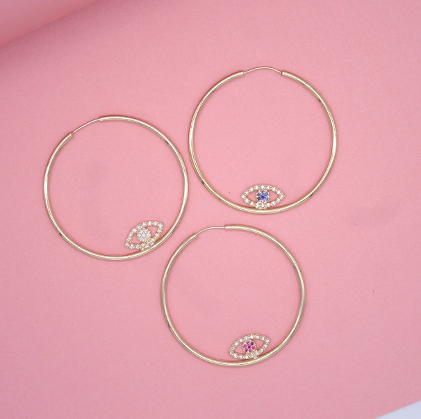 Three gold hoop earrings featuring sparkling evil eye charms rest on a soft pink background.