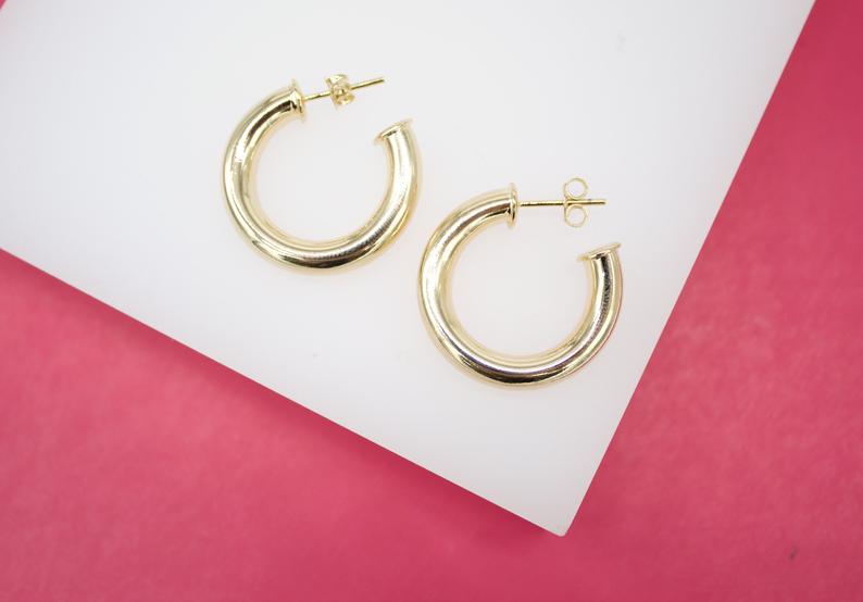 Two thick gold hoop earrings rest neatly on a white display against a vibrant pink background.