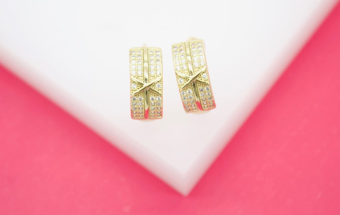 A pair of gold huggie earrings featuring sparkling cubic zirconia stones rests on a white and pink surface.