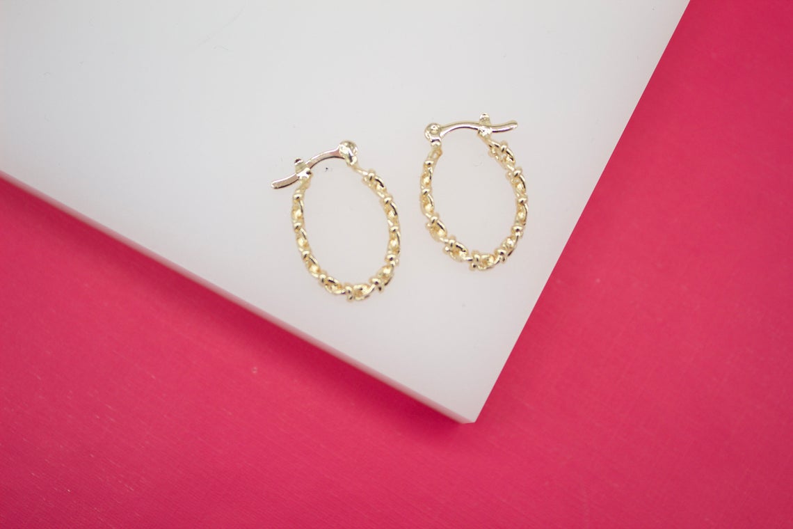 A pair of gold textured huggie hoop earrings rests on a white display against a vibrant pink background.