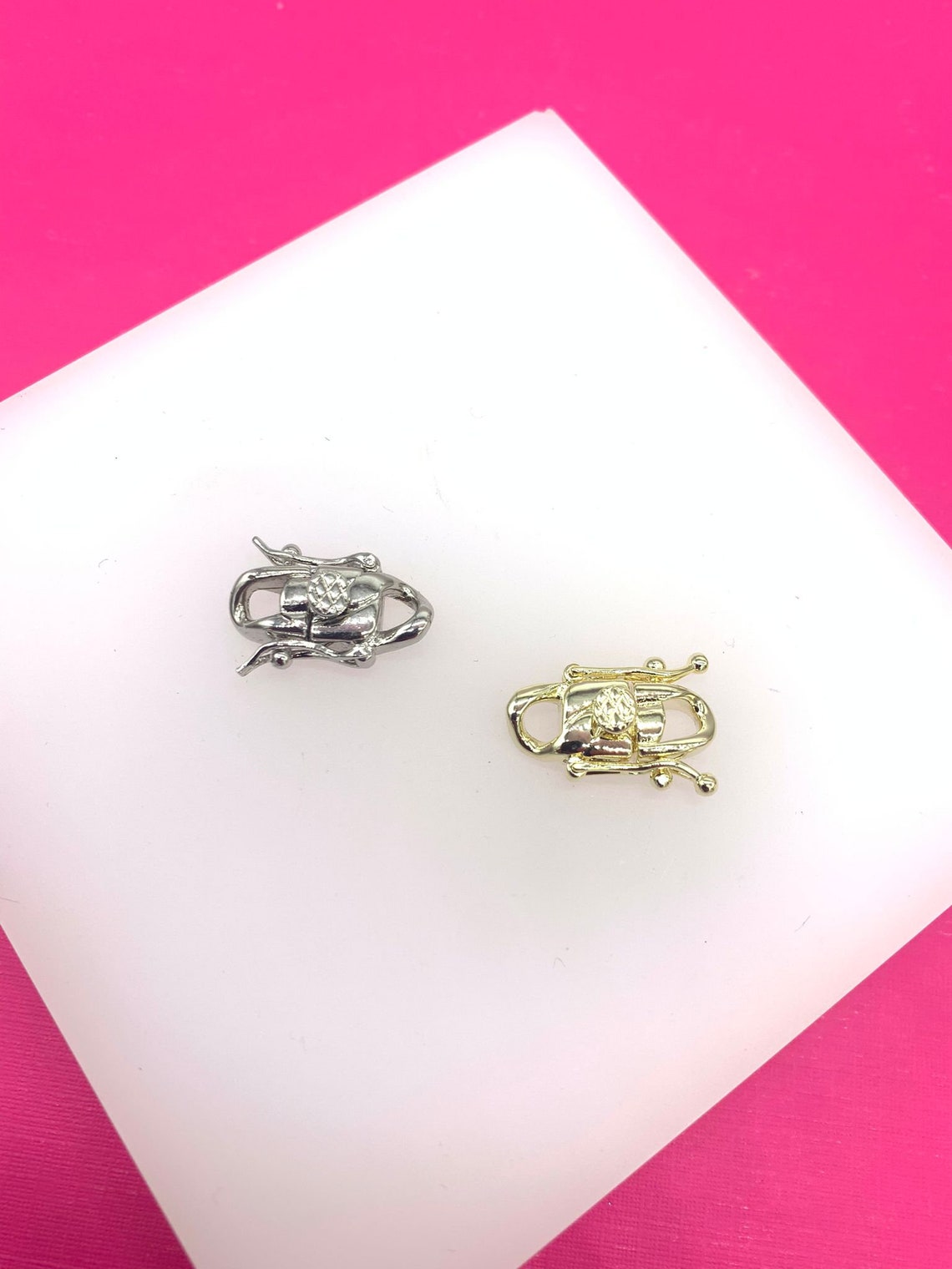 A silver fishhook clasp and a gold fishhook clasp rest side by side on a white display surface against a vibrant pink background.