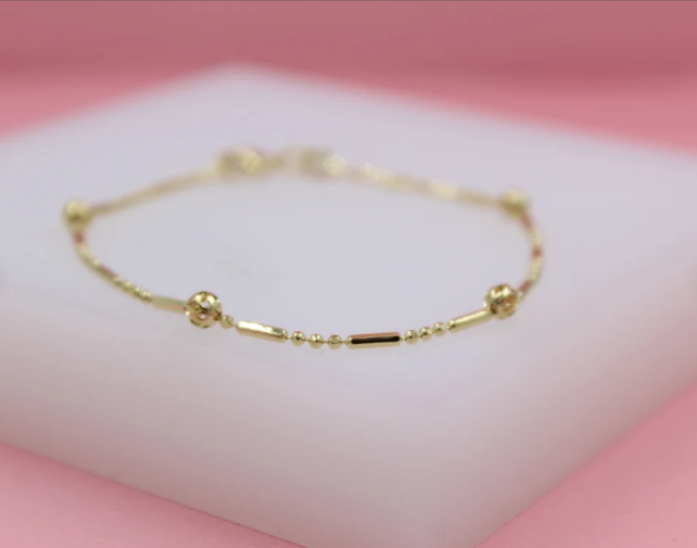 A delicate gold bracelet featuring alternating beads and cylindrical links rests on a white display stand against a soft pink background.
