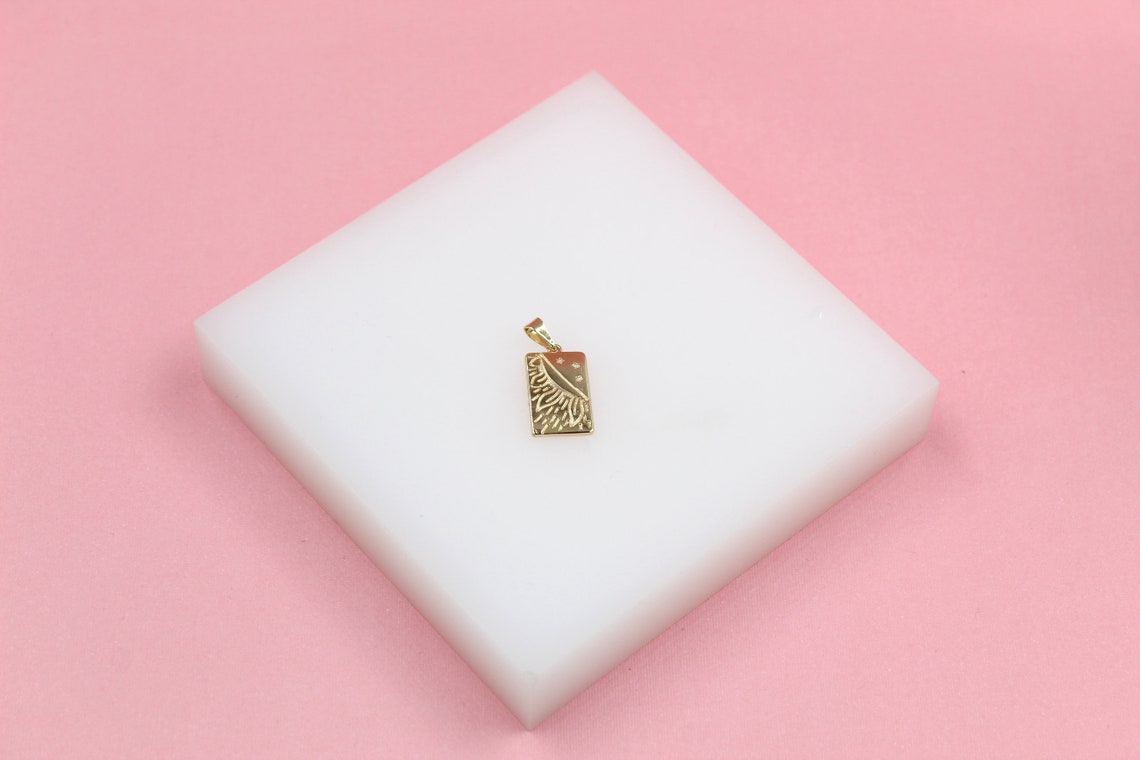 A gold celestial pendant featuring an engraved sun and moon design rests on a white display block against a pink background.