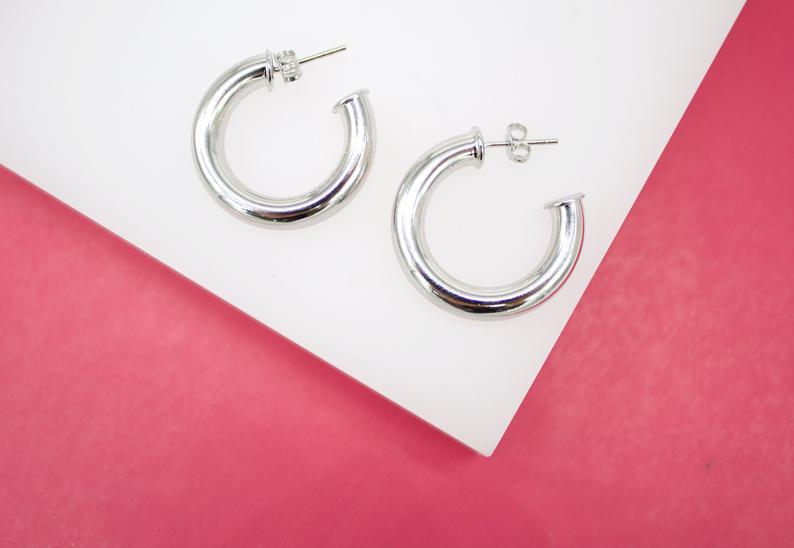A pair of thick silver hoop earrings rests on a white display surface against a vibrant pink background.