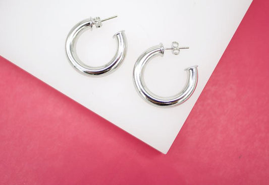 A pair of thick silver hoop earrings rests on a white display surface against a vibrant pink background.