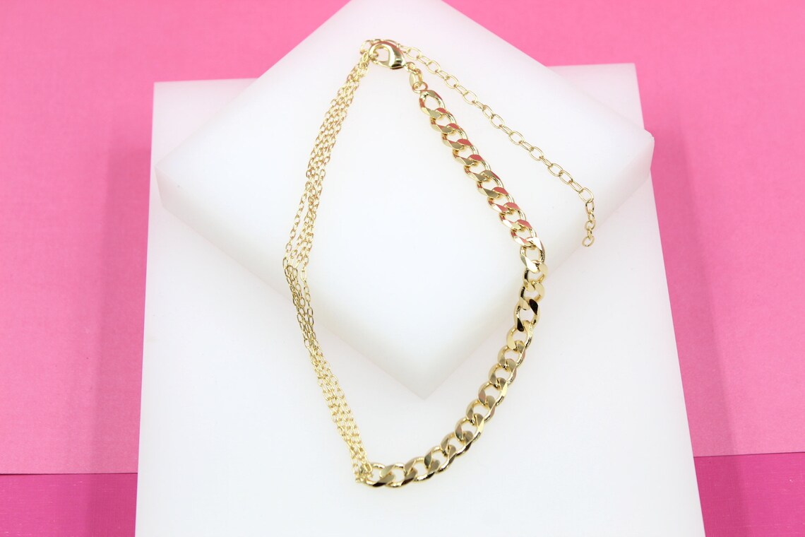 A gold-plated anklet featuring a dual-chain design rests on a white display block against a vibrant pink background.
