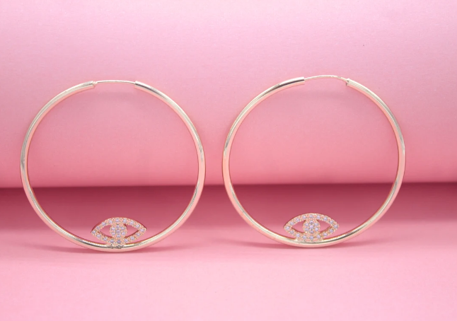 A pair of gold hoop earrings featuring sparkling evil eye charms resting at the bottom against a soft pink background.