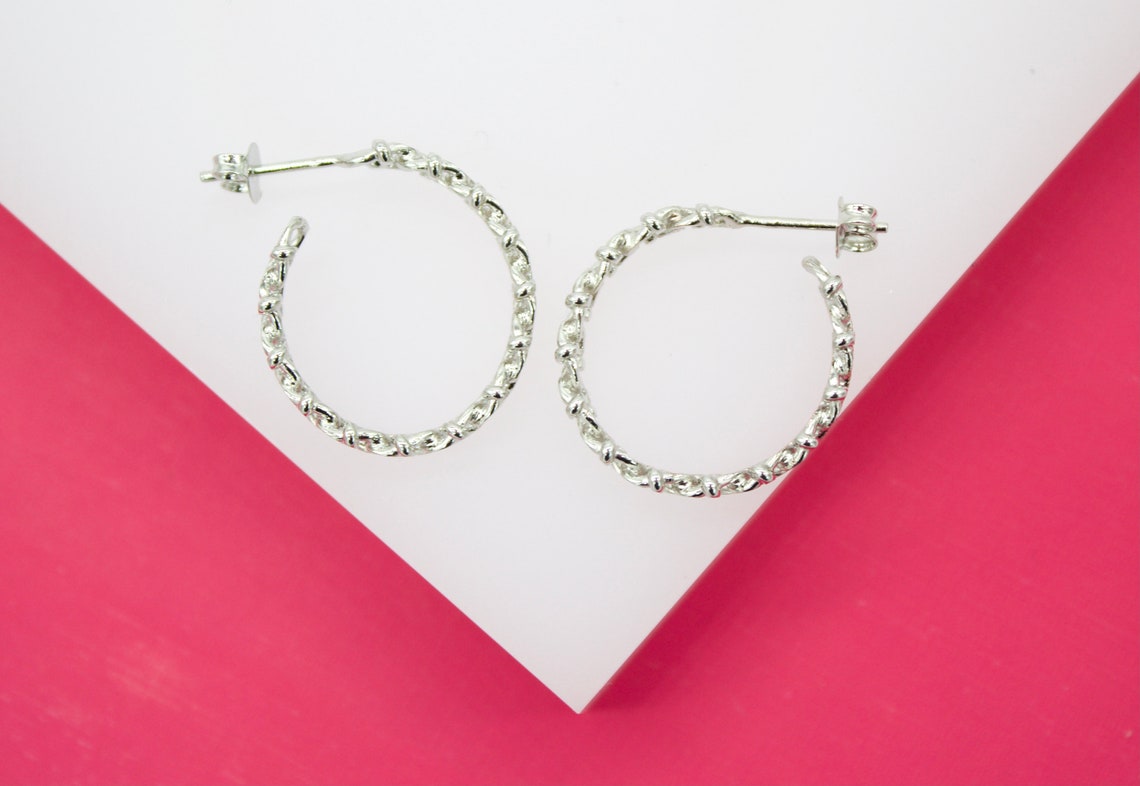 A pair of textured silver hoop earrings rests on a white display surface against a vibrant pink background.