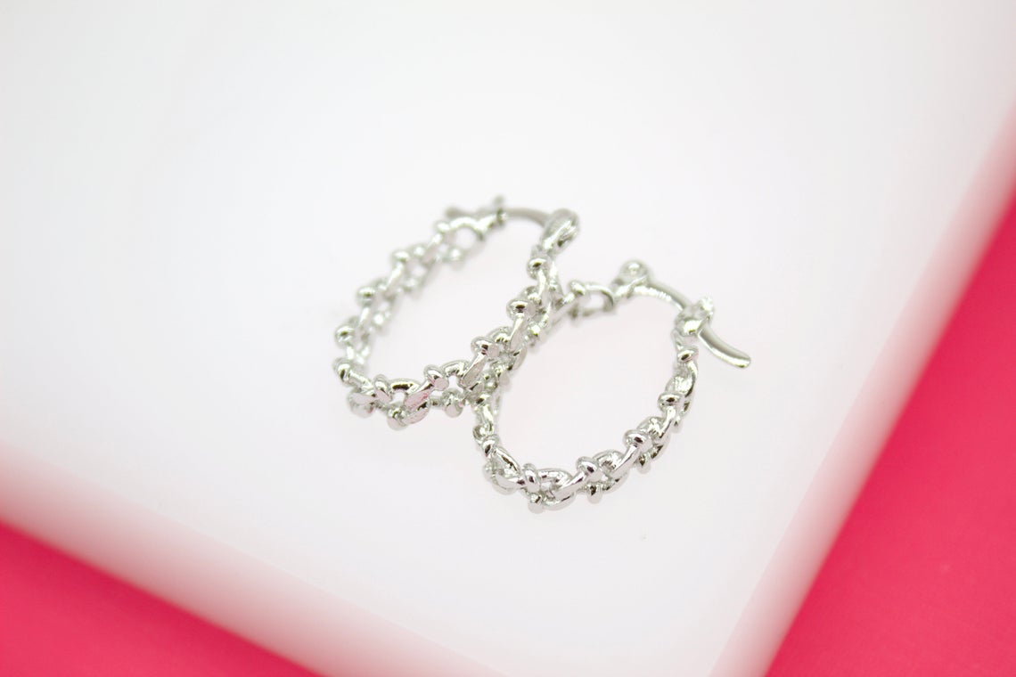 A pair of textured silver hoop earrings rests on a white display surface against a vibrant pink background.