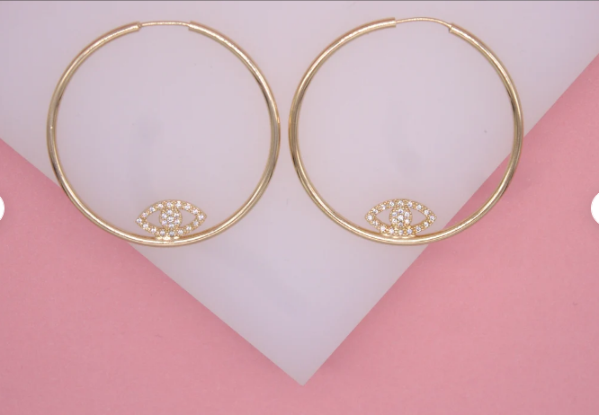 Two gold hoop earrings featuring sparkling pave diamond evil eye charms rest against a pink and white backdrop.