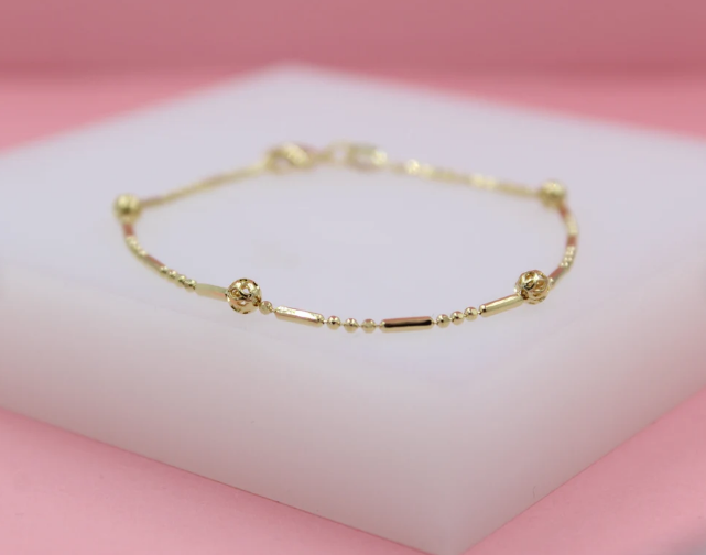A delicate gold chain bracelet featuring alternating beads and cylindrical bars rests on a white display block against a soft pink background.