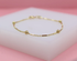 A delicate gold chain bracelet featuring alternating beads and cylindrical bars rests on a white display block against a soft pink background.