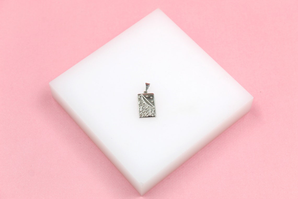 A silver rectangular pendant featuring an engraved sun and moon celestial design rests on a white display block against a pink background.