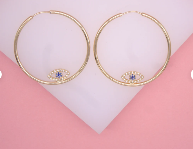 A pair of large gold hoop earrings featuring dangling evil eye charms with blue and white crystals rests on a pink and white surface.