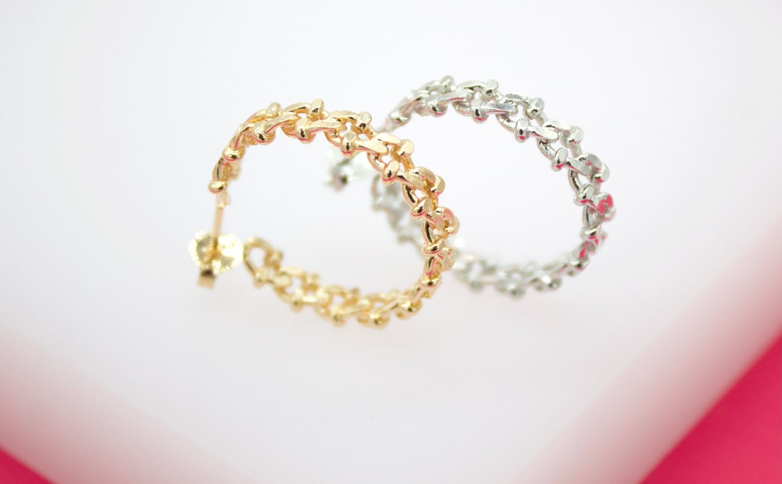 A pair of intricate, textured chain hoop earrings in gold and silver display their detailed links against a clean white and pink background.