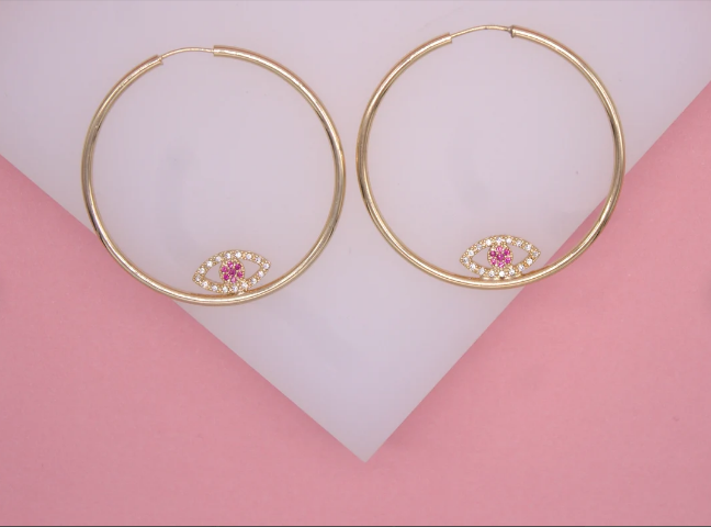 A pair of gold evil eye hoop earrings with pink and white crystal details rests on a white and pink display surface.