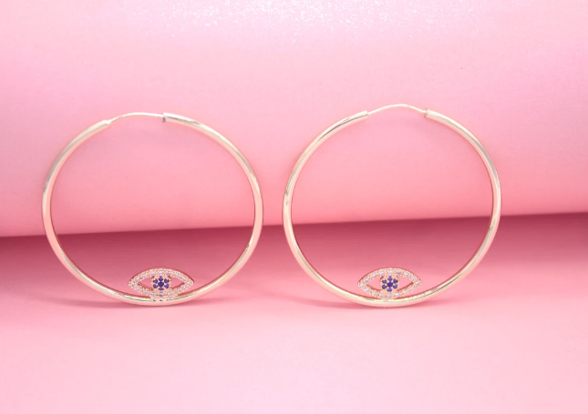 A pair of gold hoop earrings featuring a sparkling evil eye charm rests against a soft pink background.