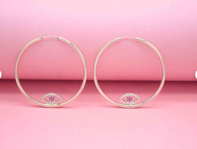 A pair of gold hoop earrings featuring sparkling evil eye charms with pink stone centers rests against a soft pink background.